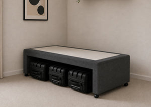 Single charcoal upholstered divan base with hidden suitcase storage, non-slip top, and black luggage underneath.