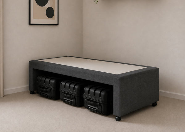 Single charcoal upholstered divan base with hidden suitcase storage, non-slip top, and black luggage underneath.