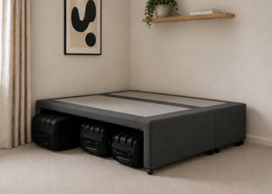 Charcoal upholstered divan base with hidden suitcase storage, non-slip top, and black luggage underneath.