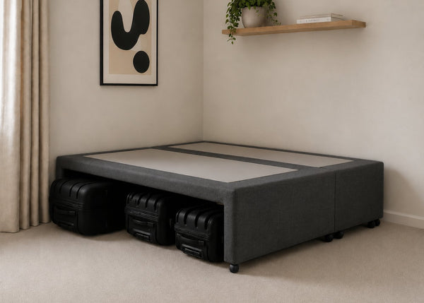 Charcoal upholstered divan base with hidden suitcase storage, non-slip top, and black luggage underneath.