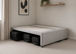 Grey upholstered divan base with hidden suitcase storage, non-slip top, and black luggage underneath.
